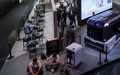 Exhibitors and visitors at a mobility trade show stand with a concept vehicle, bar tables and a large video wall, seen from above
